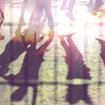 The shadows of people on a sunny concrete path, with a orange and yellow glare across the image.